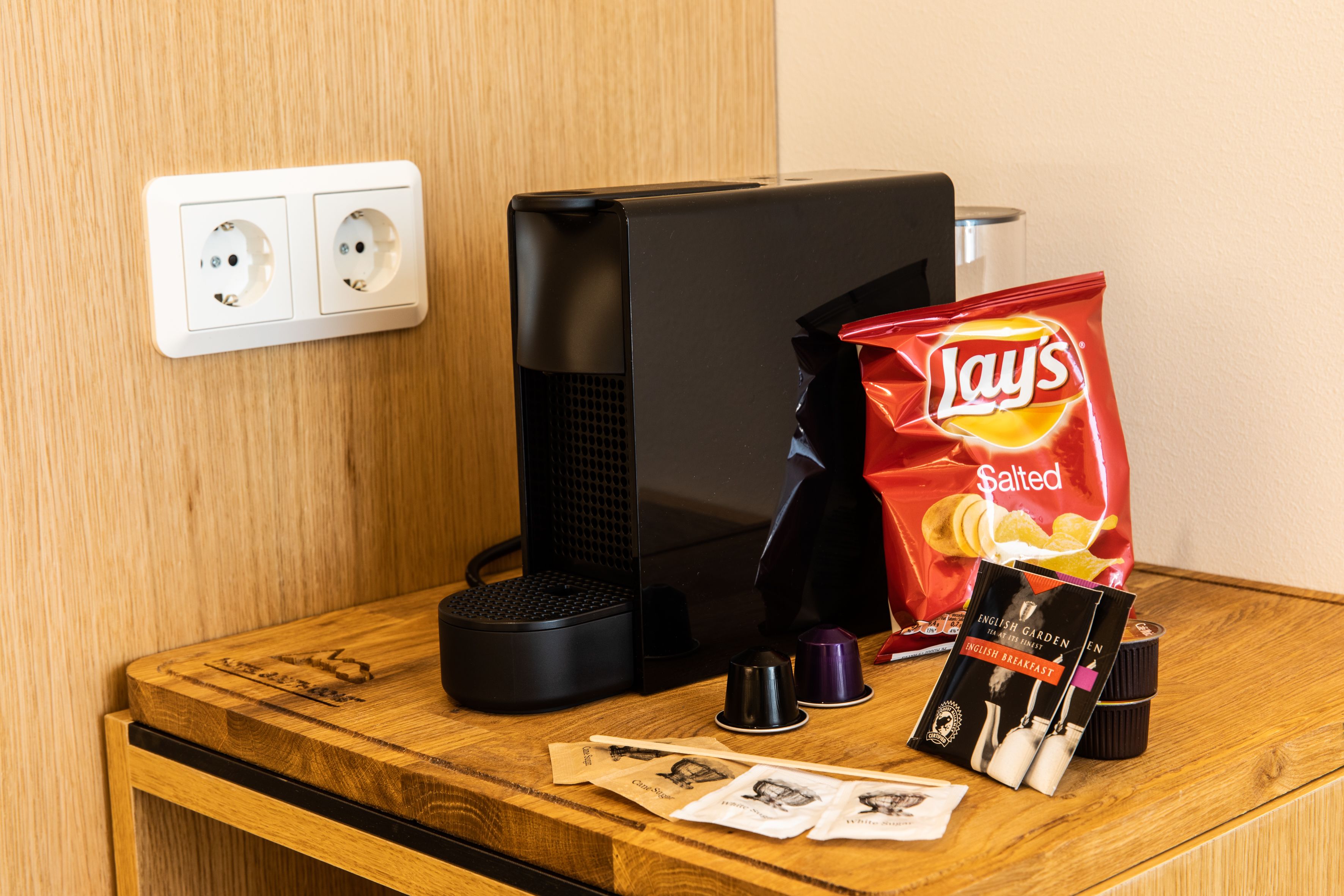 Un escritorio de madera con una computadora negra, una bolsa roja de papas Lay's y algunas tazas de café.