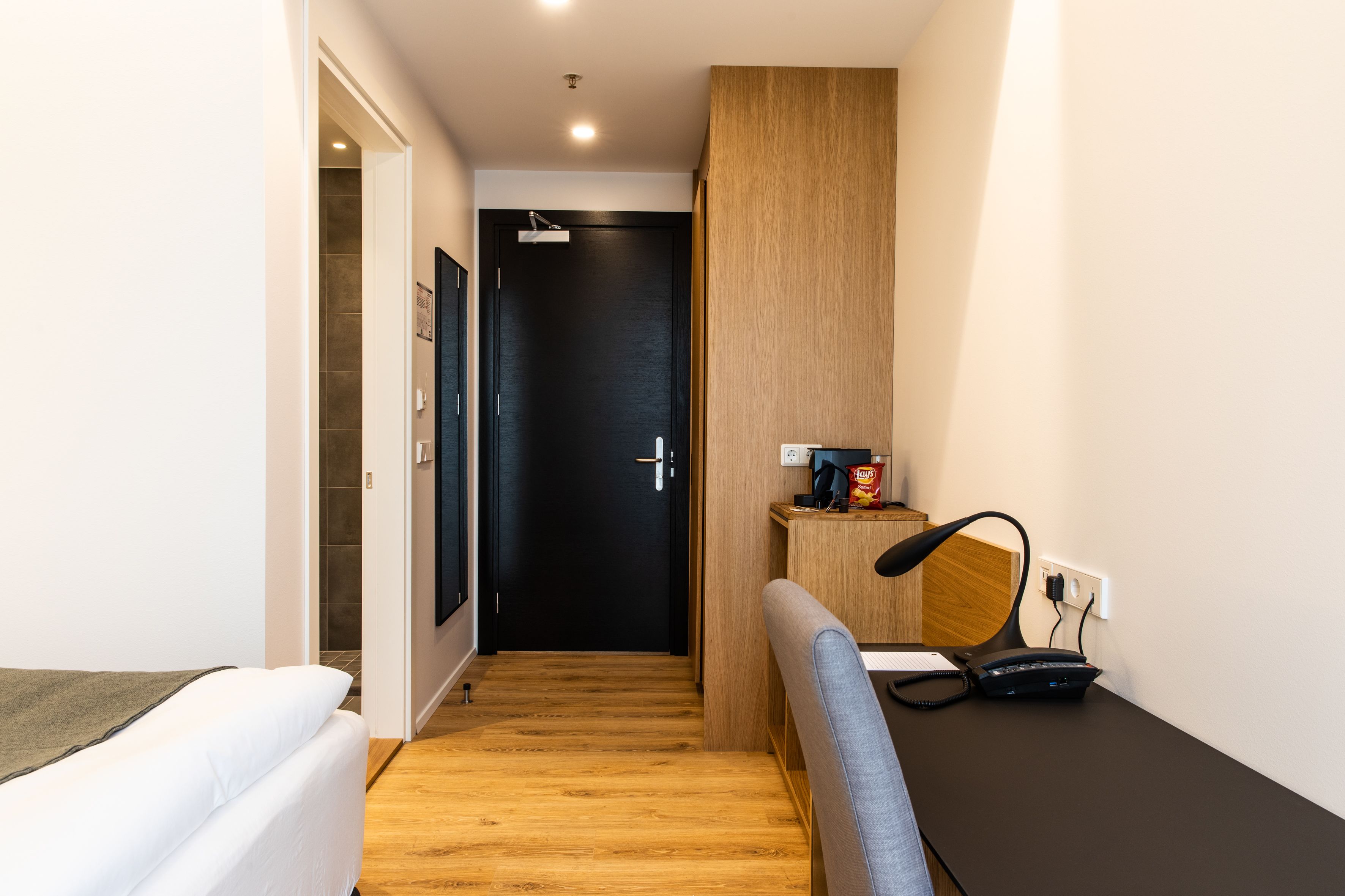 A narrow hallway with a bed on the left, a desk with a chair and a phone on the right, and a door at the end.