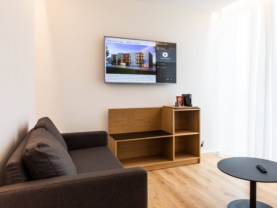 Una habitación de hotel moderna con un televisor de pantalla plana grande mostrando un interior de hotel, una estantería de madera, un sofá marrón y una mesita redonda pequeña.