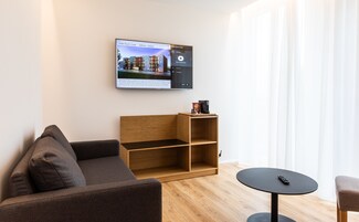 Una habitación de hotel moderna con un televisor de pantalla plana grande mostrando un interior de hotel, una estantería de madera, un sofá marrón y una mesita redonda pequeña.
