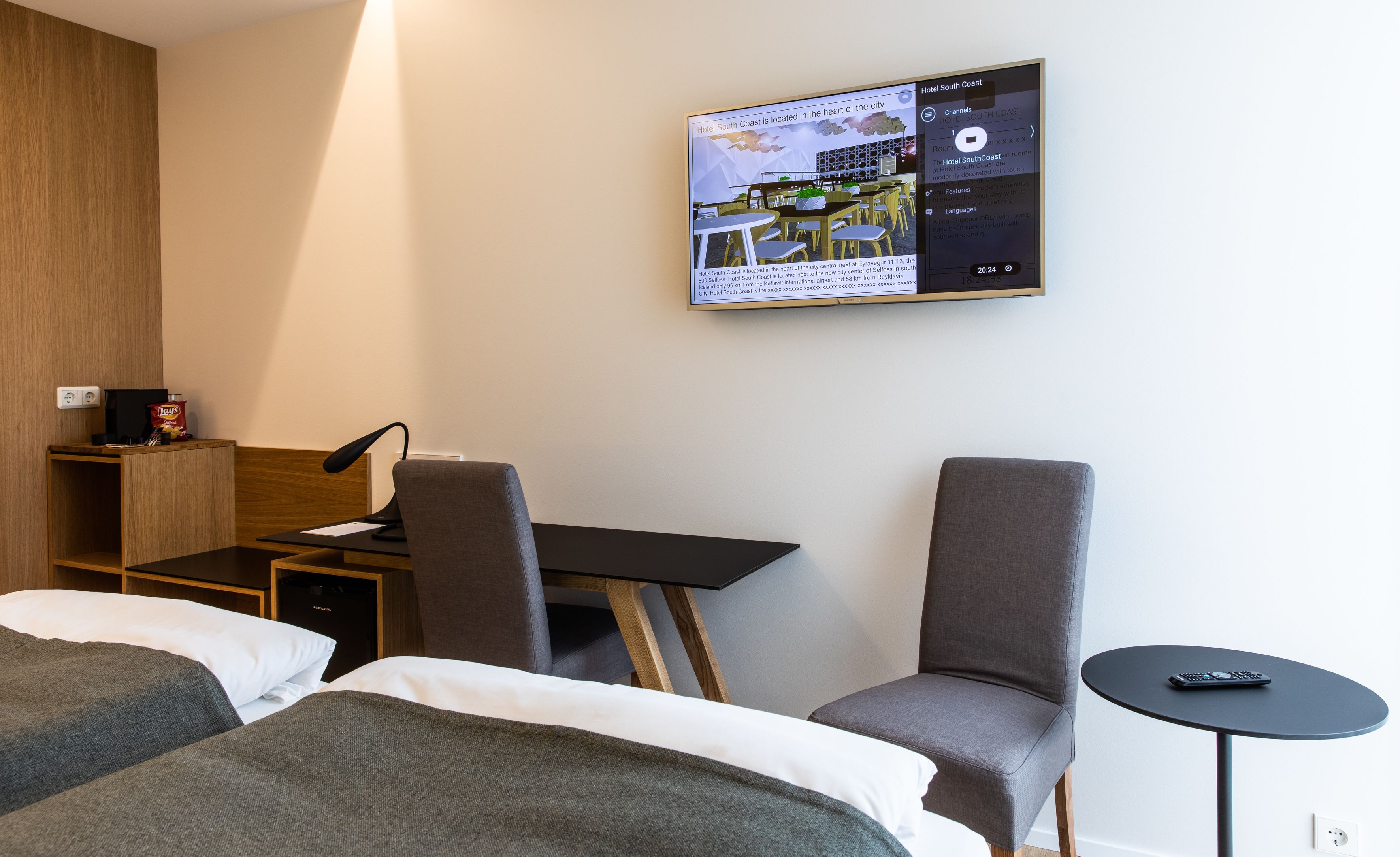 A hotel room with two beds, a desk, and a chair. There is a television mounted on the wall displaying hotel information.