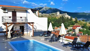 A pool area with lounge chairs and umbrellas, a building with a balcony, and a mountainous backdrop. - Holiday House Above with pool (Klis)