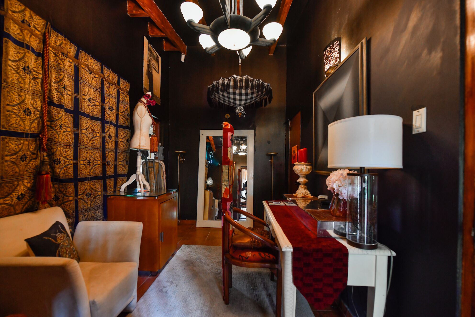 Un pasillo con papel tapiz estampado, un sillón blanco, una silla de madera, un mantelino rojo a cuadros y una lámpara blanca.