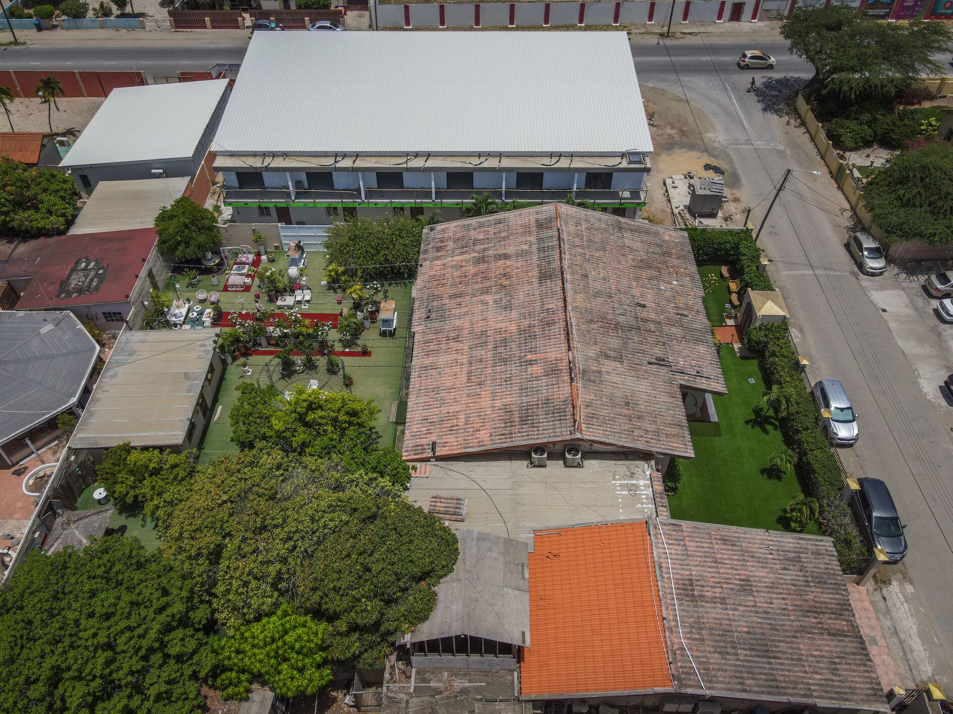 An aerial view of a building with a large flat roof, surrounded by greenery and a parking area.