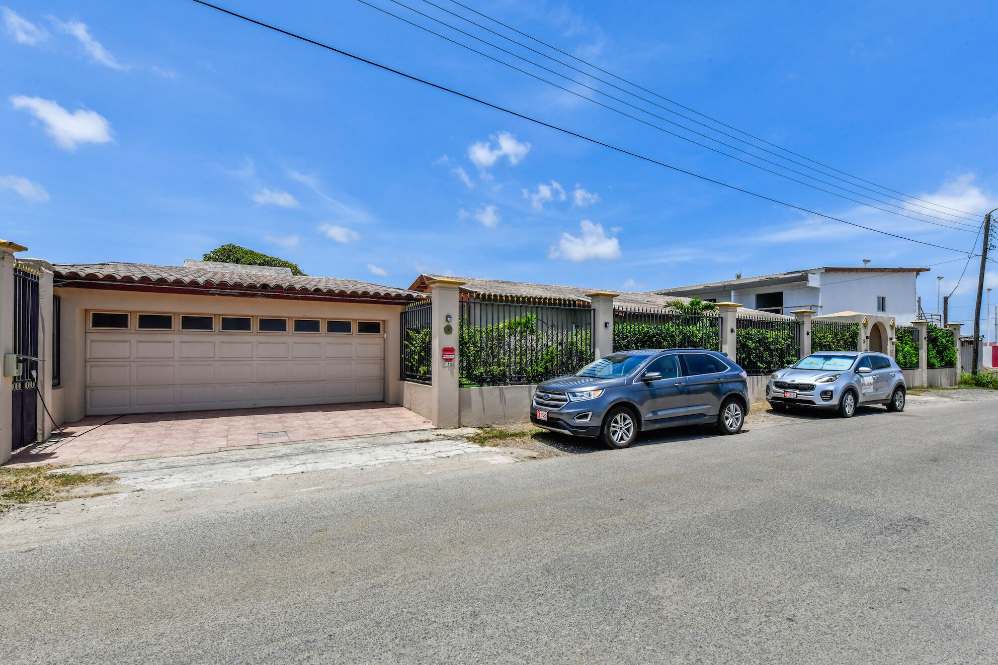 A residential street with two parked cars, a garage, and a gated entrance.