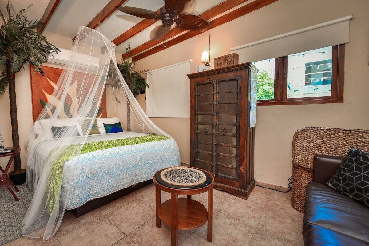 A bedroom with a bed under a canopy, a wooden door, a wicker sofa, and a small round table.