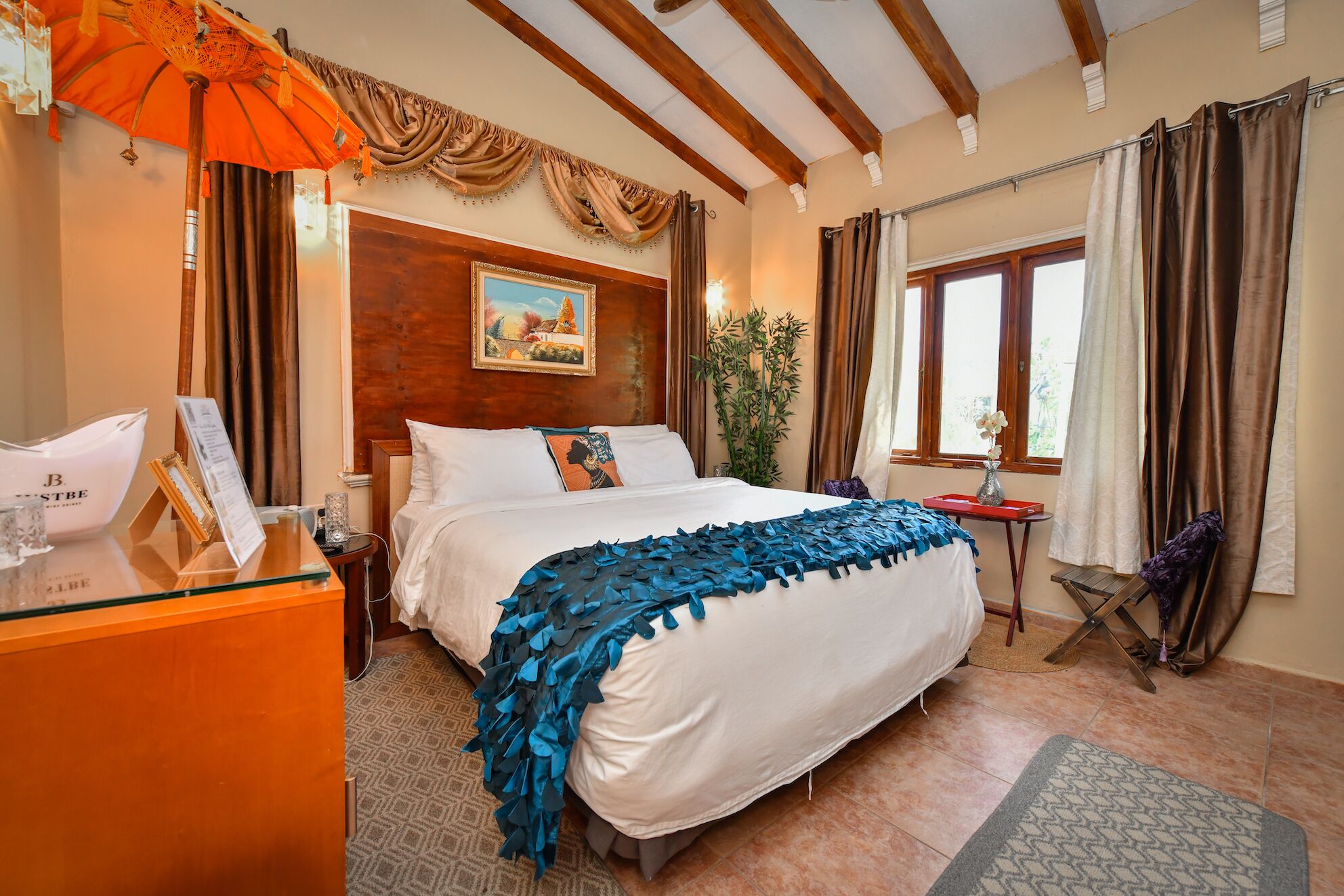 Habitación de hotel con una cama grande, vigas de madera en el techo, una ventana con cortinas, una mesita con un florero y una planta.