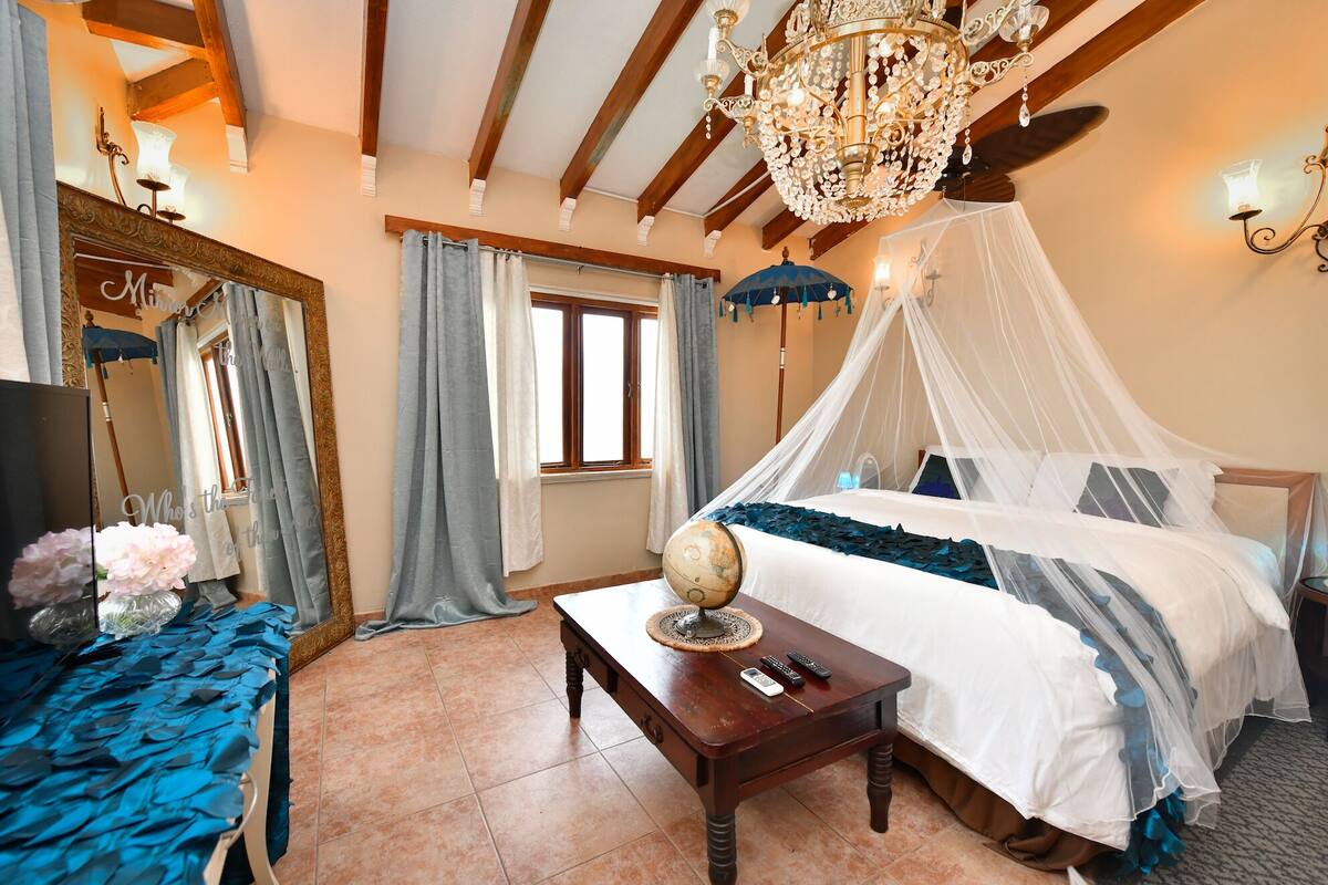 A bedroom with a four-poster bed, a chandelier, a globe on the nightstand, and a large mirror.