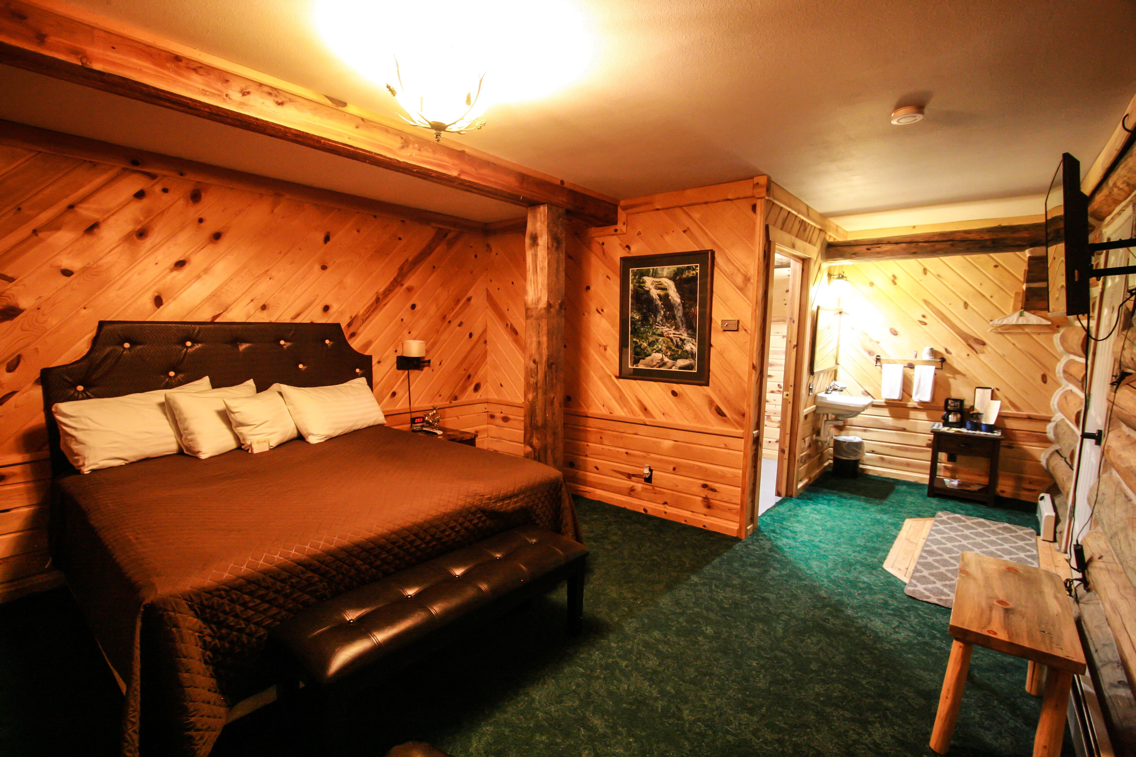 A wooden cabin bedroom with a bed, bench, dresser, and a small table with a lamp.