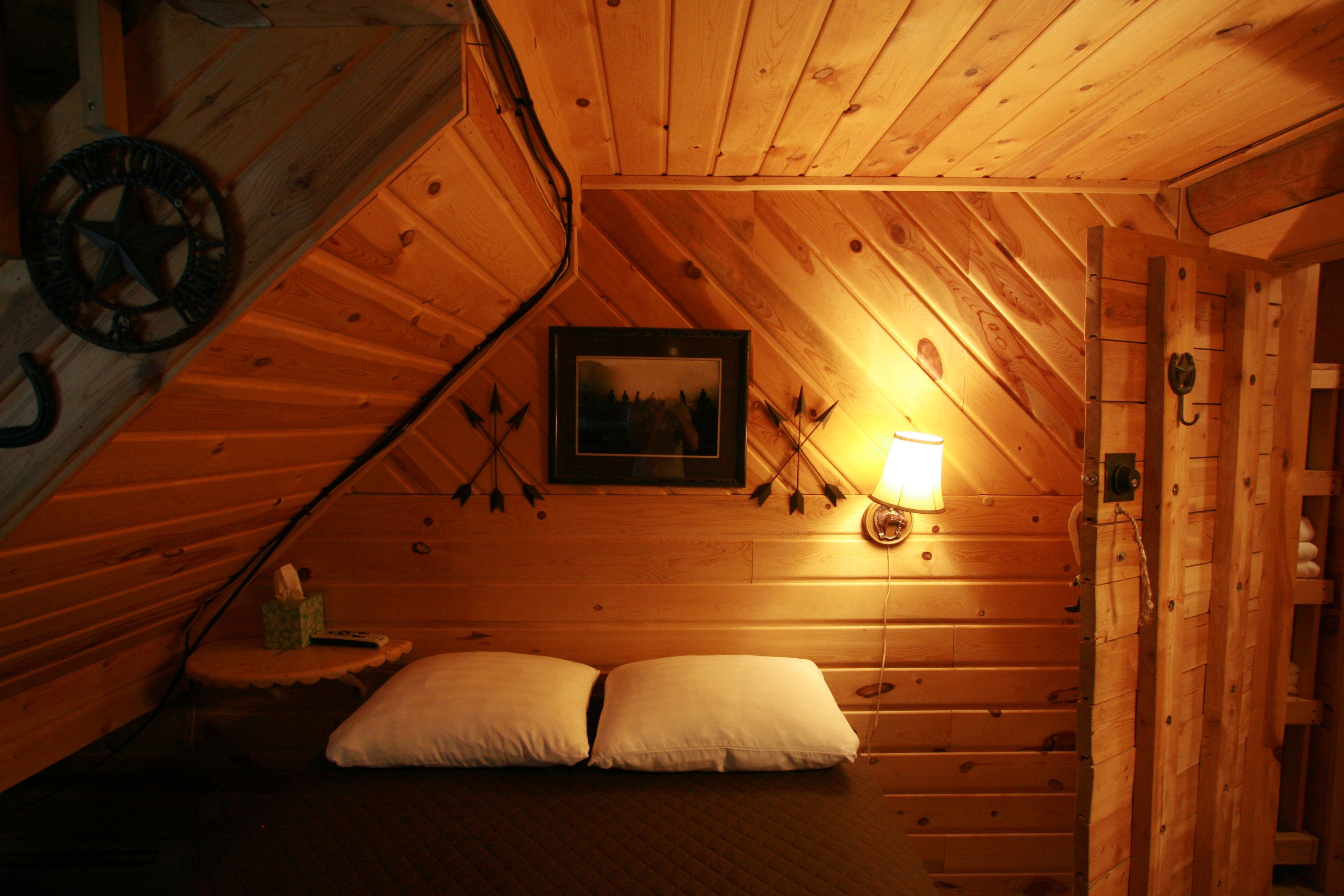 A cozy attic bedroom with wooden walls, a framed picture, a bedside lamp, and two pillows.