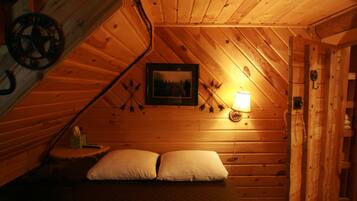 A cozy attic bedroom with wooden walls, a framed picture, a bedside lamp, and two pillows.