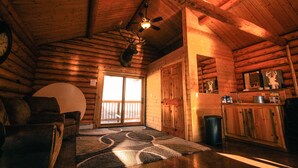 A log cabin interior with a ceiling fan, a fireplace, a dining area, and a large window. - Judith Mountain Lodge (Lewistown)