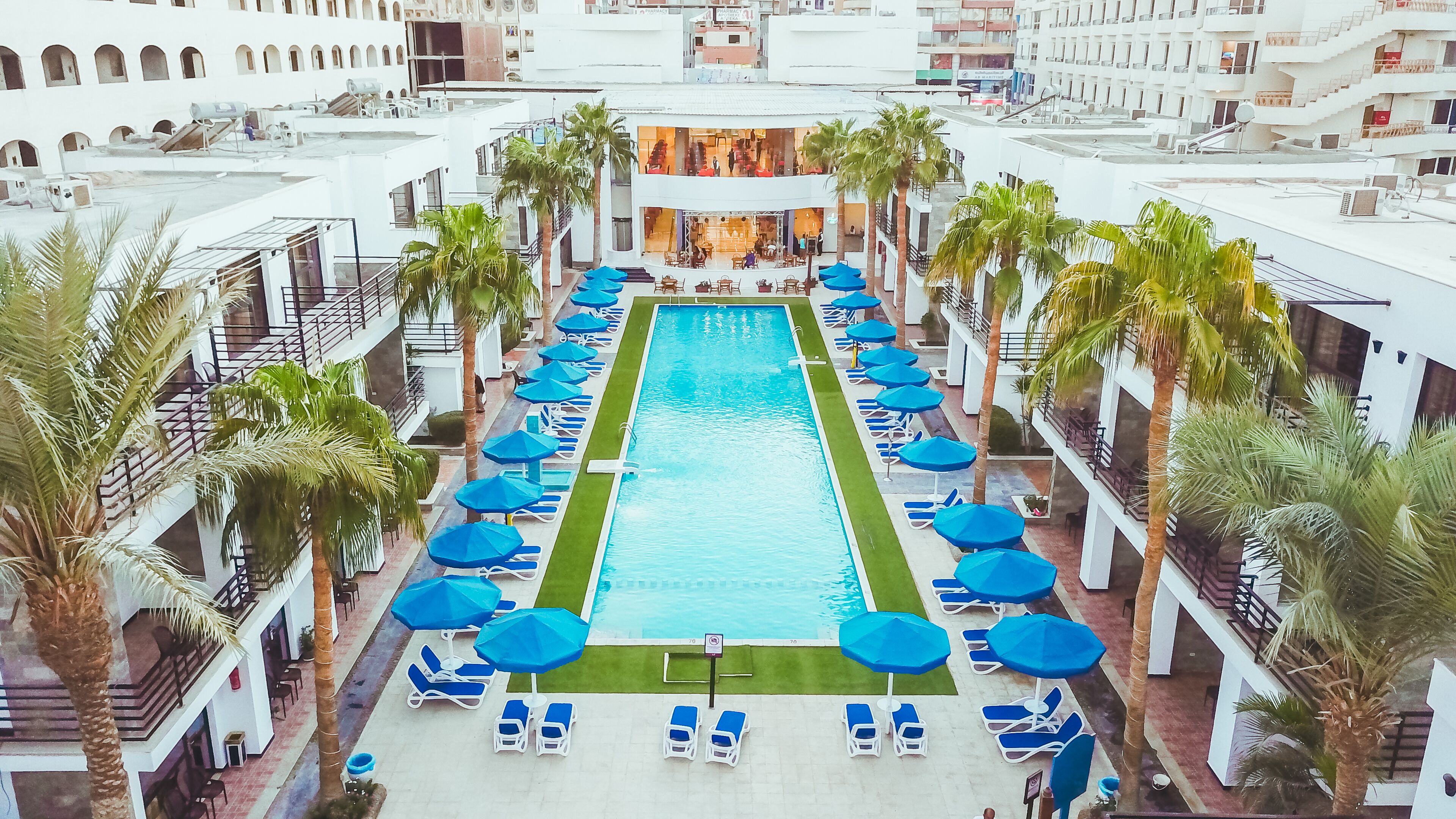 Ein Swimmingpool mit blauen Sonnenschirmen und Liegestühlen, umgeben von Palmen und einem mehrstöckigen Gebäude mit Balkonen.