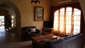 A living room with a sofa, a coffee table, a TV on a wooden stand, a painting on the wall, and a large window with curtains. - Ta Cikku, a luxury 5 bedroom with villa  with pool for family, friends and fun.  (Gharb)