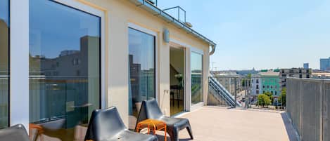 A rooftop terrace with two black plastic chairs and a small orange table, overlooking a cityscape with buildings and a staircase.