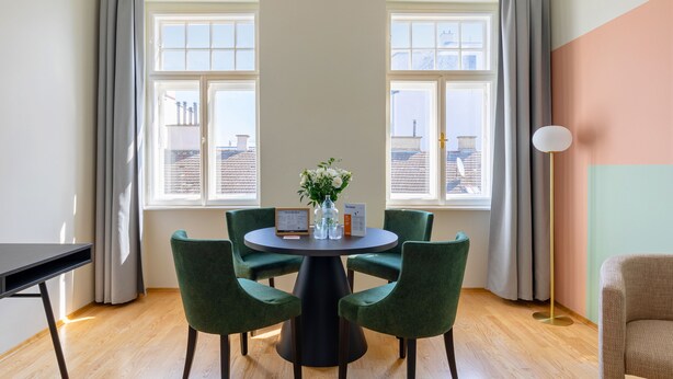 Een kamer met een ronde tafel, vier groene stoelen, een vaas met bloemen en een ingelijst certificaat.