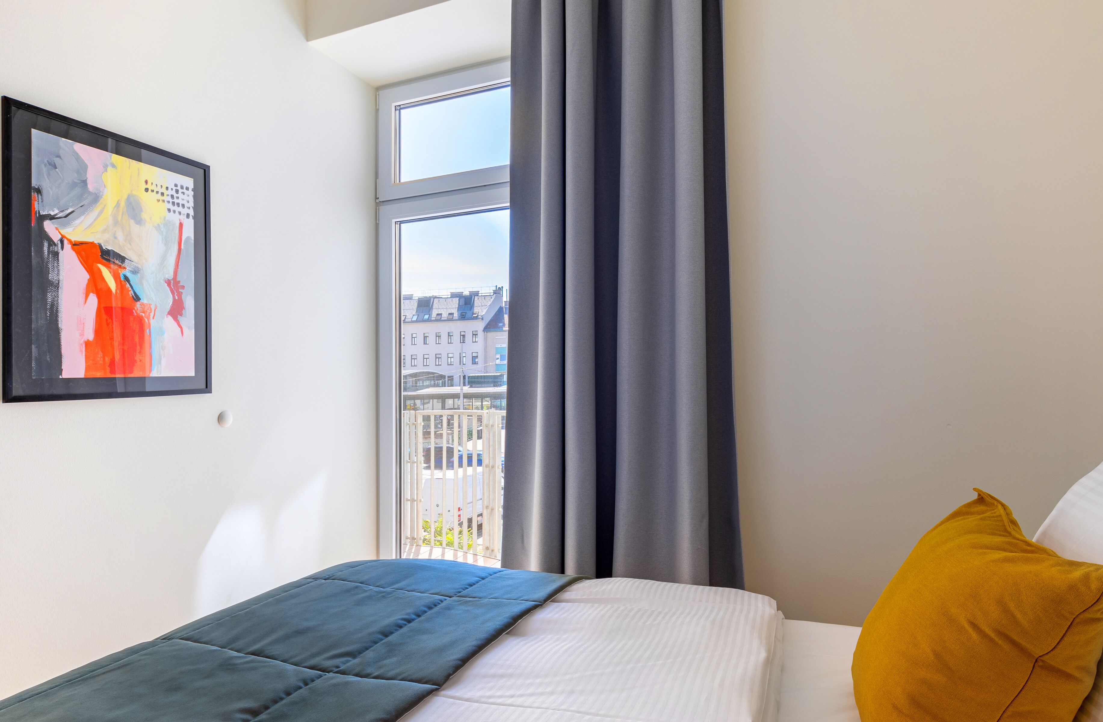Quarto com cama, janela com cortinas, vista para a cidade e um quadro abstrato emoldurado na parede.