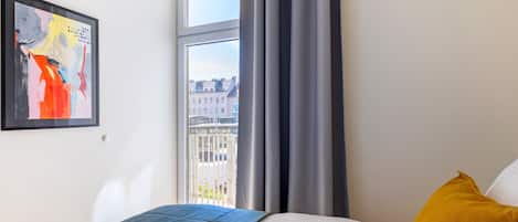 A bedroom with a bed, a window with curtains, a city view, and a framed abstract painting on the wall.