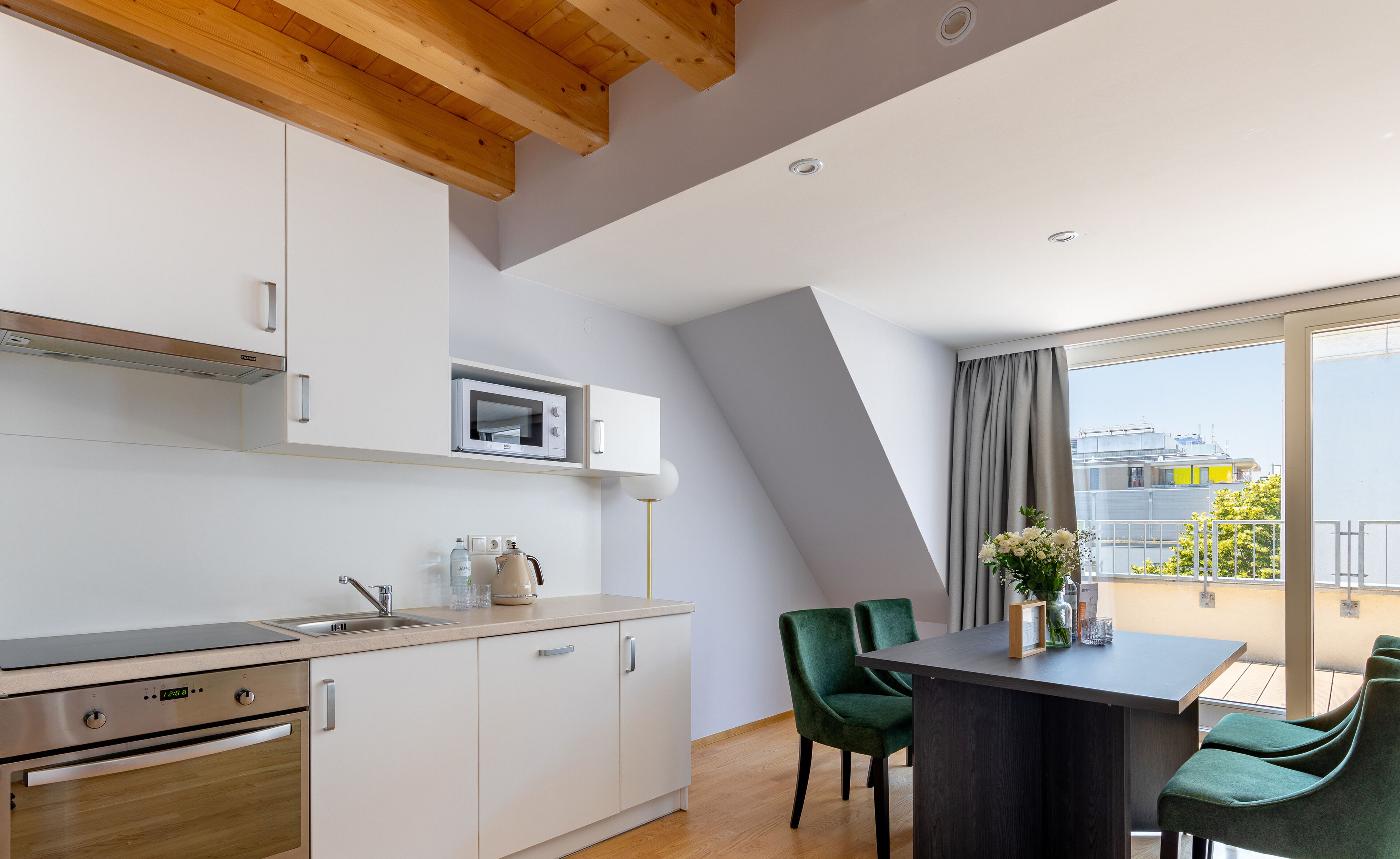 Cocina moderna con armarios blancos, microondas y estufa de acero inoxidable. Cuenta con una zona de comedor con mesa y sillas, y un balcón con vistas a edificios.