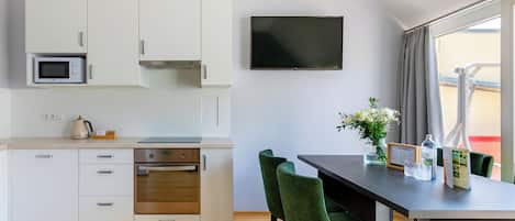 Een moderne keuken met witte kasten, een magnetron, een fornuis en een flatscreen televisie. Er staat een eettafel met groene stoelen en een bloemenvaas.