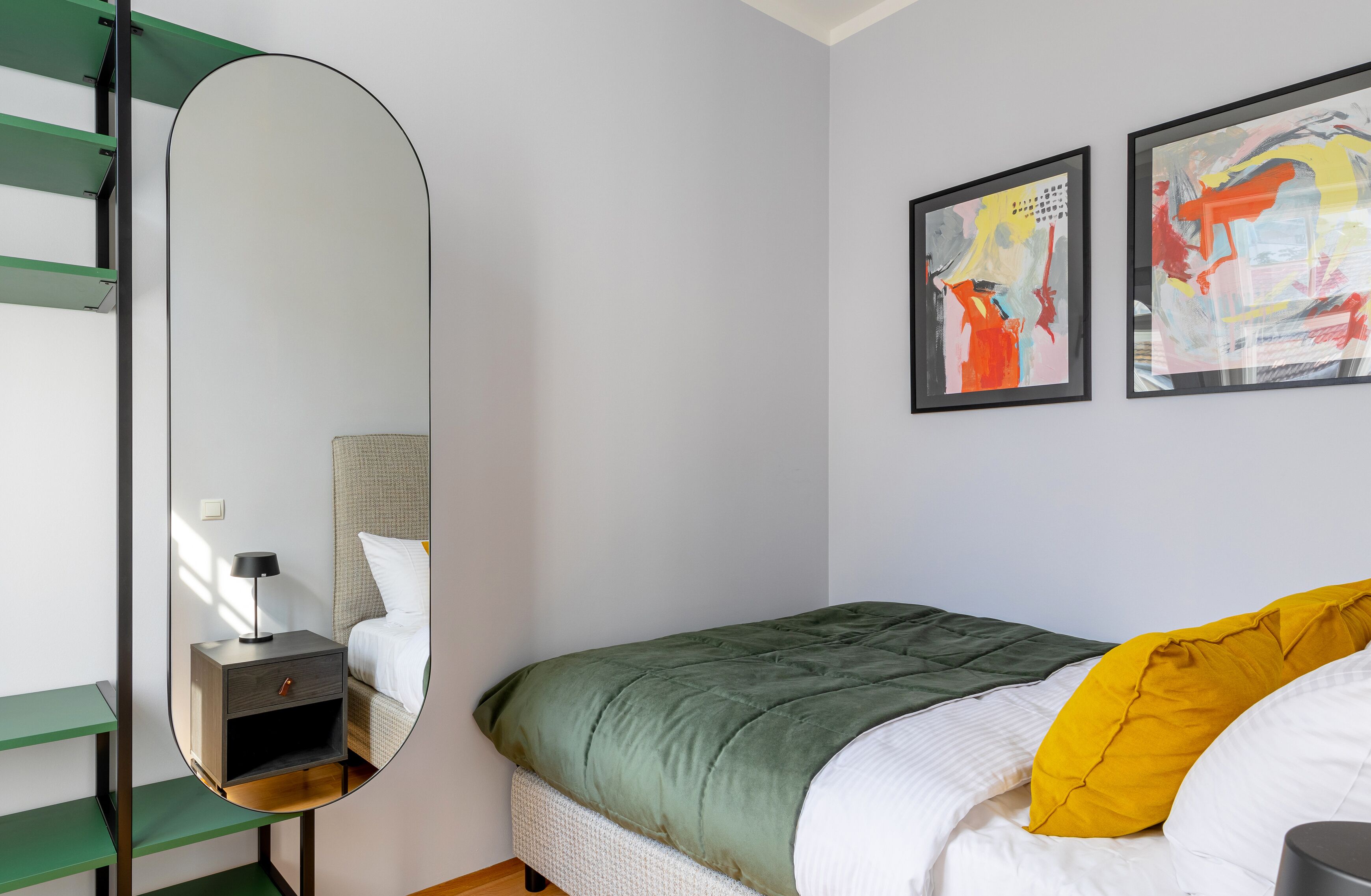Een slaapkamer met een bed, een nachtkastje met een lamp, een spiegel en twee abstracte schilderijen aan de muur.