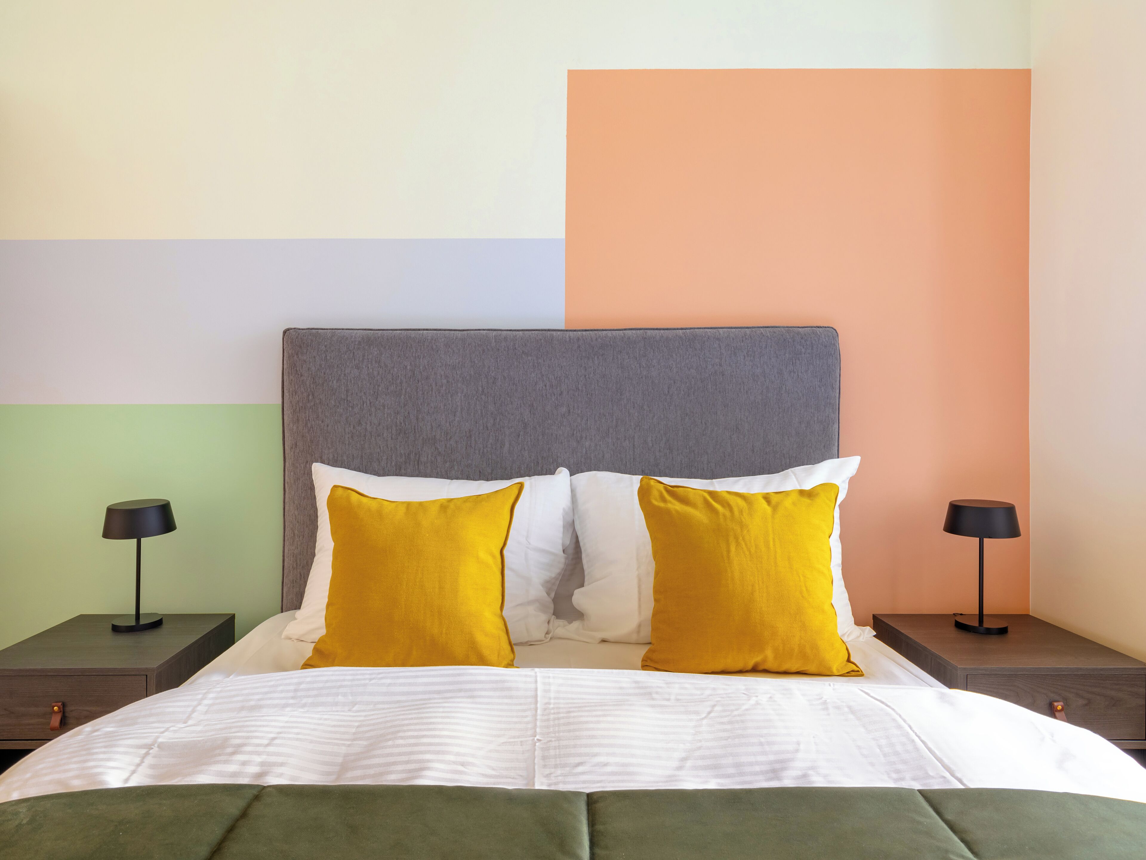 A bed with a grey headboard, white and yellow pillows, and a green bedspread. There are two bedside tables with black lamps.