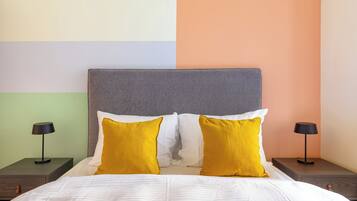 A bed with a grey headboard, white and yellow pillows, and a green bedspread. There are two bedside tables with black lamps.