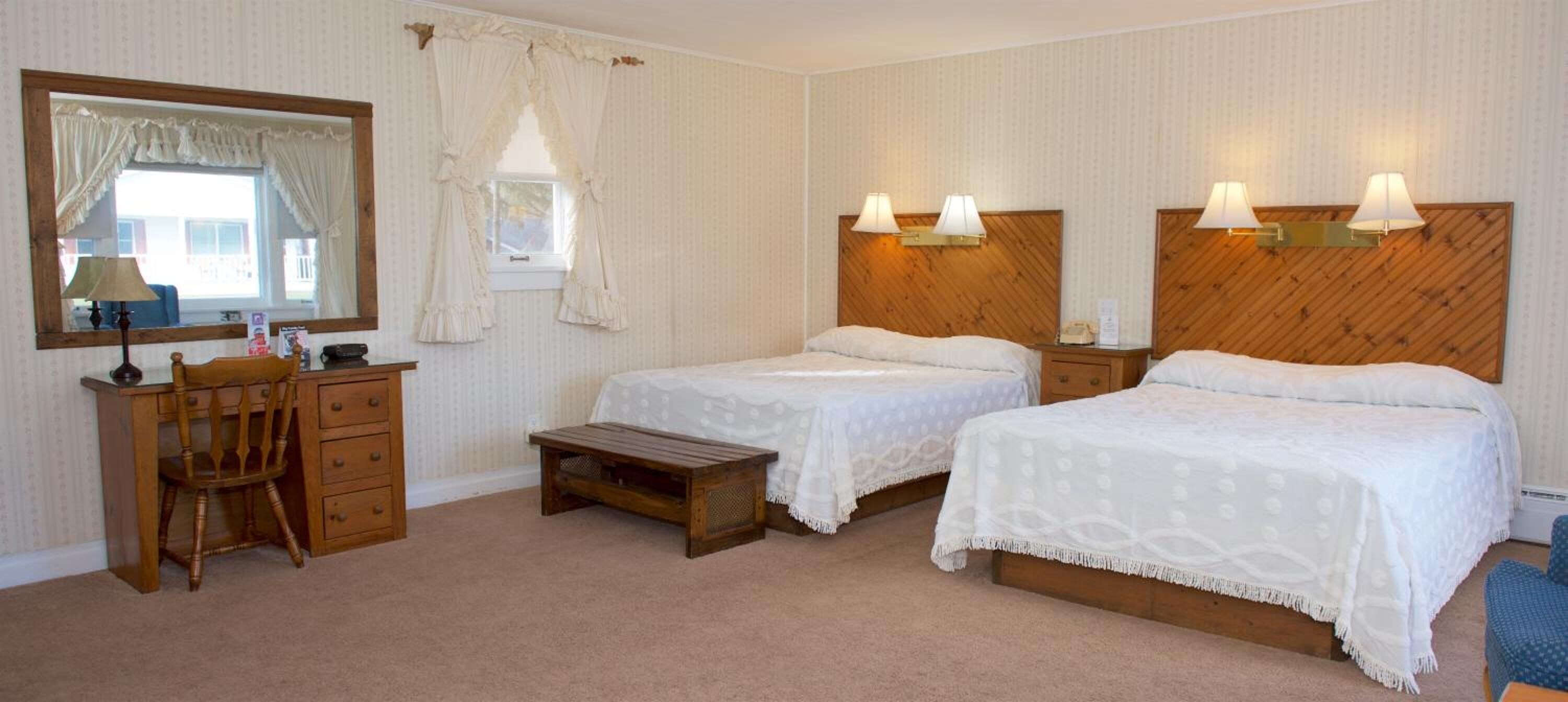 Une chambre d’hôtel avec deux lits, une tête de lit en bois, un bureau avec une chaise, un petit banc en bois et un miroir.
