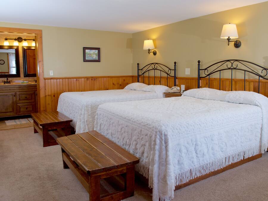 A hotel room with two beds, a wooden bench, a vanity with a mirror, and wall-mounted lamps.