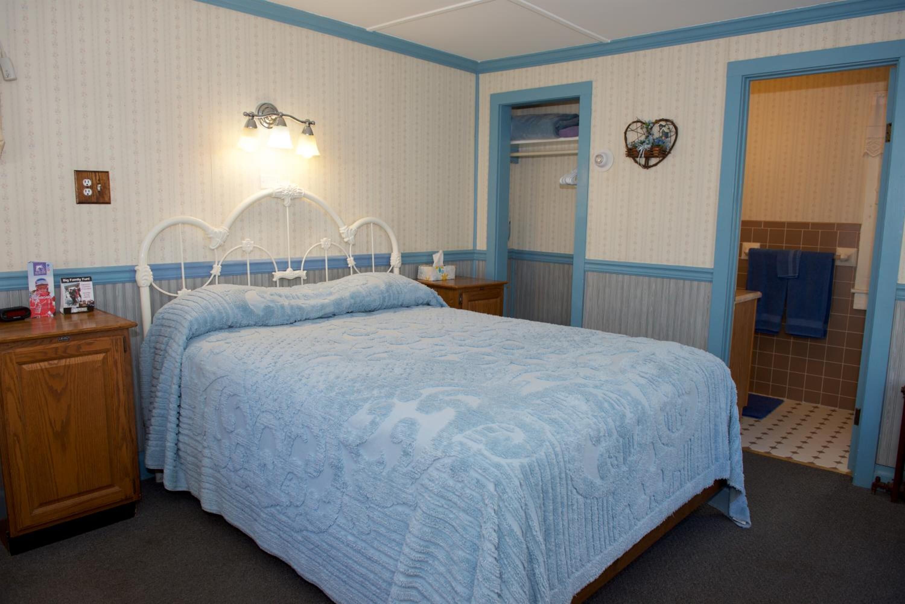 Quarto com cama, mesas de cabeceira, luminária na parede, prateleira pequena e porta do banheiro.