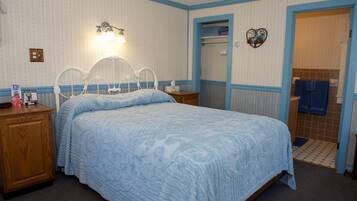 Quarto com cama, mesas de cabeceira, luminária na parede, prateleira pequena e porta do banheiro.