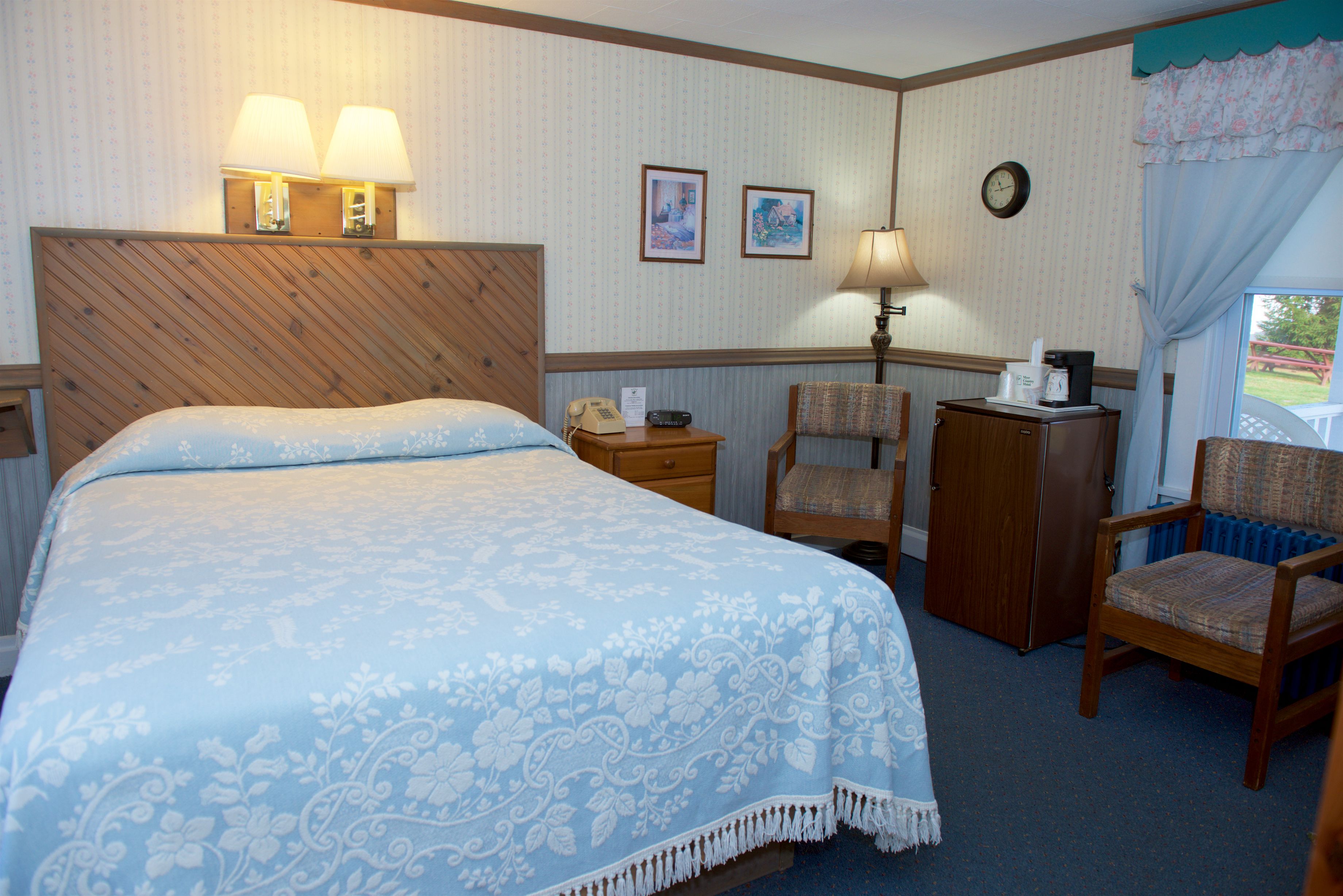 A hotel room with a bed, two chairs, a desk with a telephone, a clock, and framed pictures on the wall.
