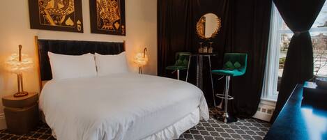 A hotel room with a large bed, two wall-mounted lamps, a green chair, a mirror, and a view of the outside.