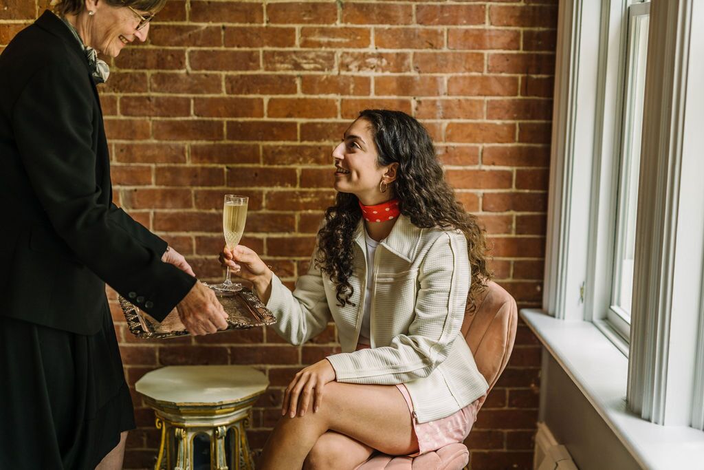A person holding a tray with a glass of champagne, sitting on a stool in front of a brick wall.