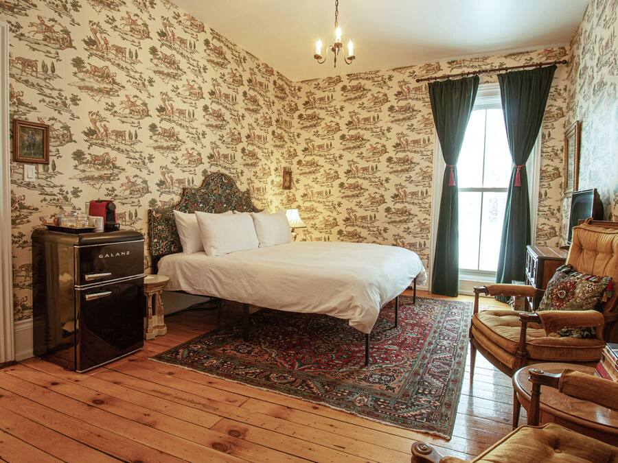 Ein Zimmer mit einem Bett, einem alten Galanz-Kühlschrank, Holzboden, einem gemusterten Teppich und einem Sessel mit Büchern.
