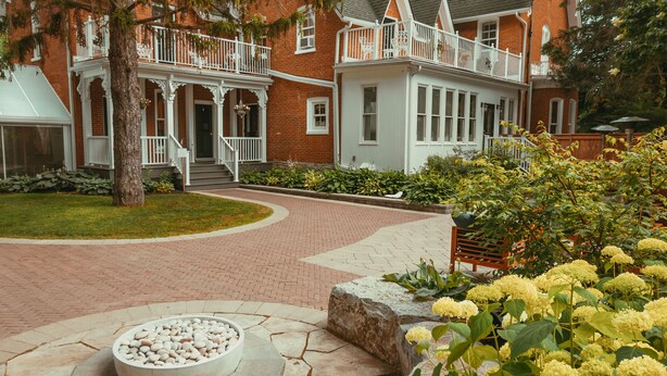 A large brick house with a wrap-around porch, a stone fire pit, and a garden with yellow flowers. - Merrill House (Prince Edward)