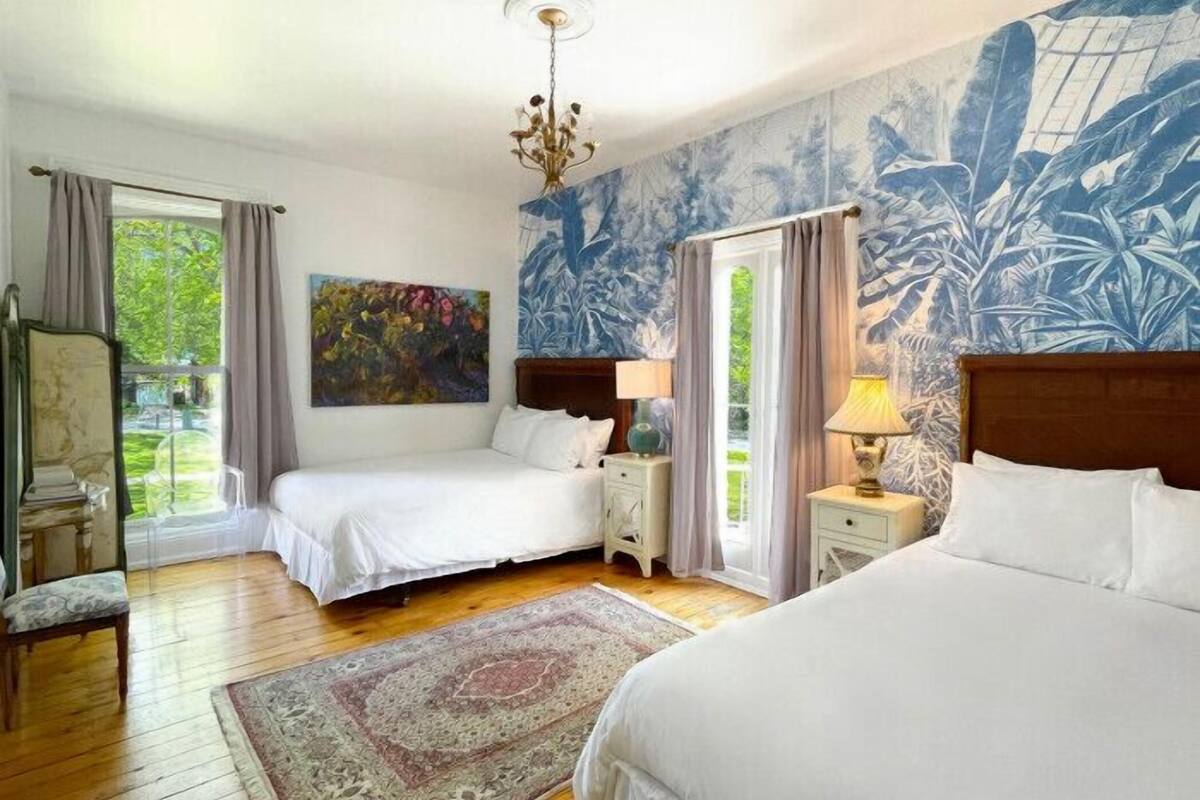 A bedroom with two beds, a large window, a chandelier, and a tropical mural.