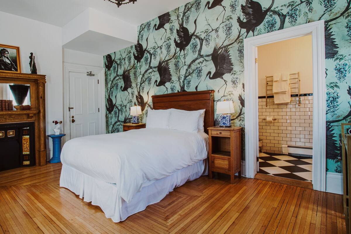 A bedroom with a large bed, wooden floor, a dresser with a mirror, and a wall with a bird mural.
