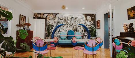 A room with a blue sofa, pink and blue chairs, a large abstract painting, and a piano.