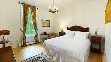A bedroom with a large bed, a chandelier, a window with curtains, a bedside table, and a chair.