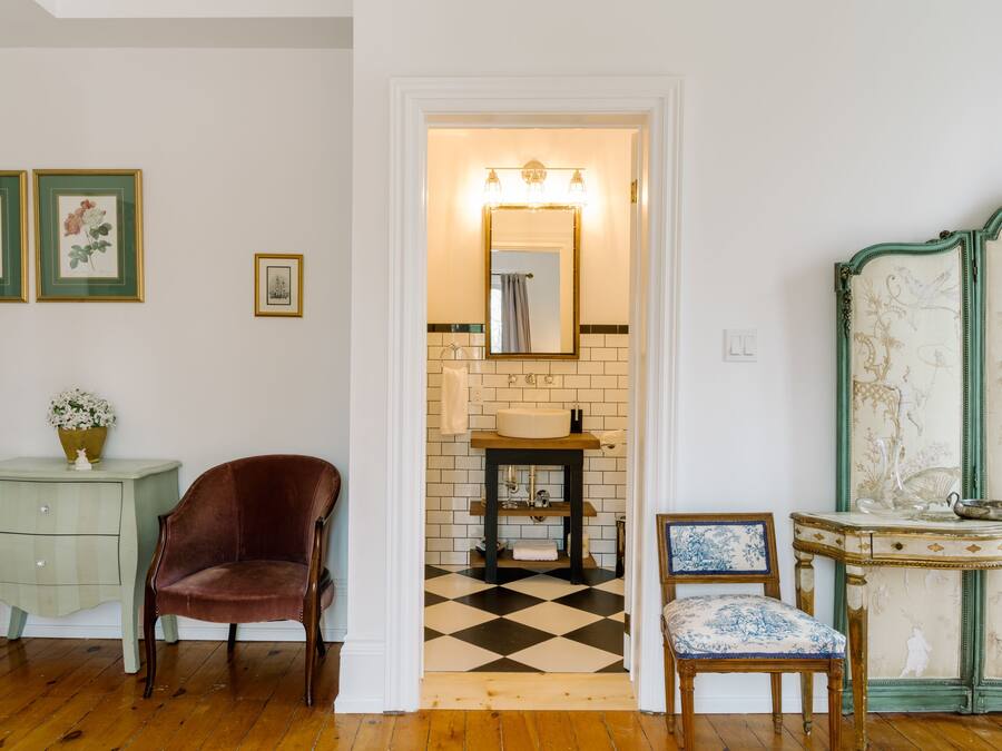 A room with a checkered floor, a vintage chair, a small table with a vase, and a mirror with a light above it.
