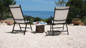 Dos sillas frente a una mesa con una bandeja de objetos, sobre un suelo pedregoso con vista al mar y árboles al fondo.
