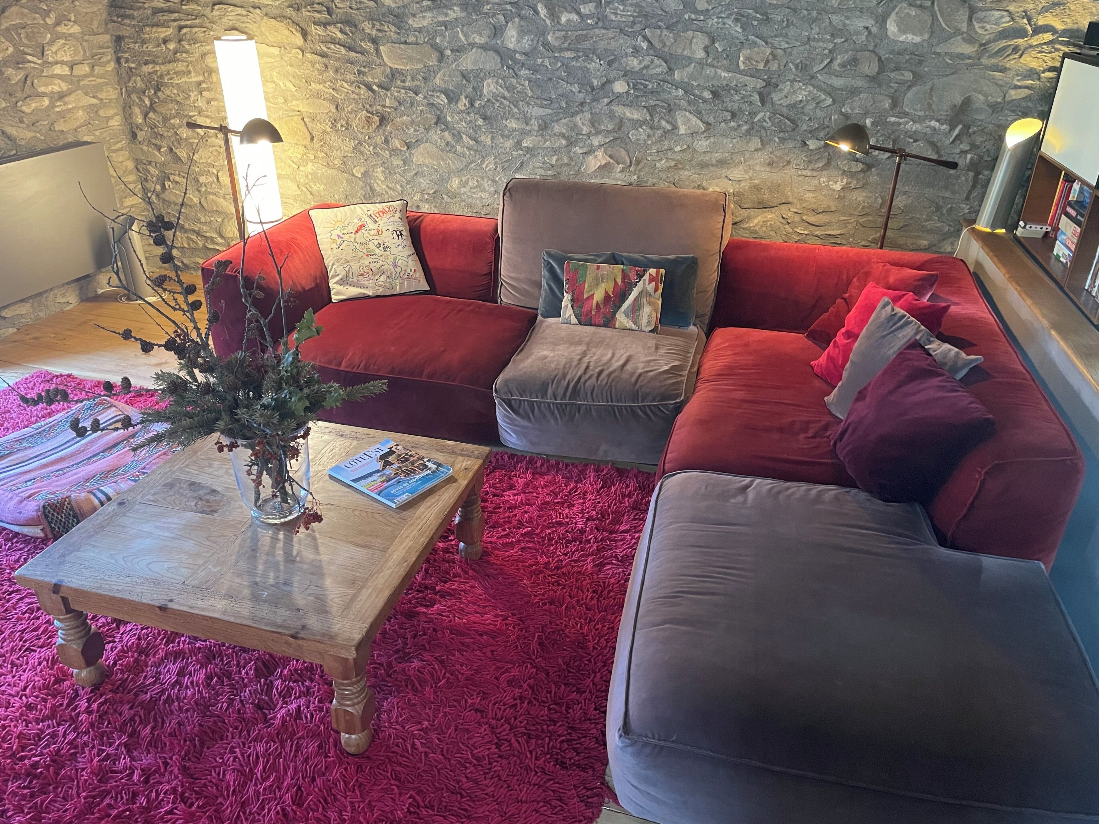 A cozy living room with a stone wall, a red and grey sectional sofa, a wooden coffee table, and a red shag rug.