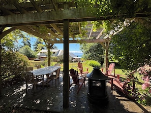 En overdækket terrasse med et bord og stole, et bålsted og udsigt til huse og bjerge.