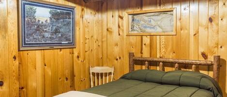 A wooden cabin room with a bed, a framed picture on the wall, and a small chair.