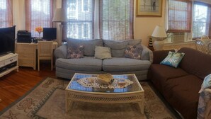 Uma sala de estar com um sofá cinzento, um sofá castanho, uma mesa de centro de vidro com objetos decorativos, uma televisão e uma pequena mesa com um abajur.