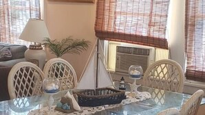 Uma área de refeições com uma mesa de vidro cercada por cadeiras de vime, um quadro na parede e uma planta em vaso.