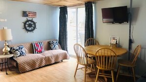 Una sala de estar acogedora con techo de madera, un sofá con almohadones de temática náutica, una mesita con una lámpara y un comedor con sillas de madera.