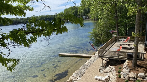 Un lago tranquilo con un muelle de madera que se adentra en el agua, rodeado de un muro de piedra y una terraza con mobiliario exterior.