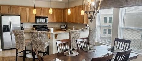 A modern kitchen with wooden cabinets, a stainless steel refrigerator, a microwave, and a coffee maker. There are wicker chairs around a wooden dining table with placemats.